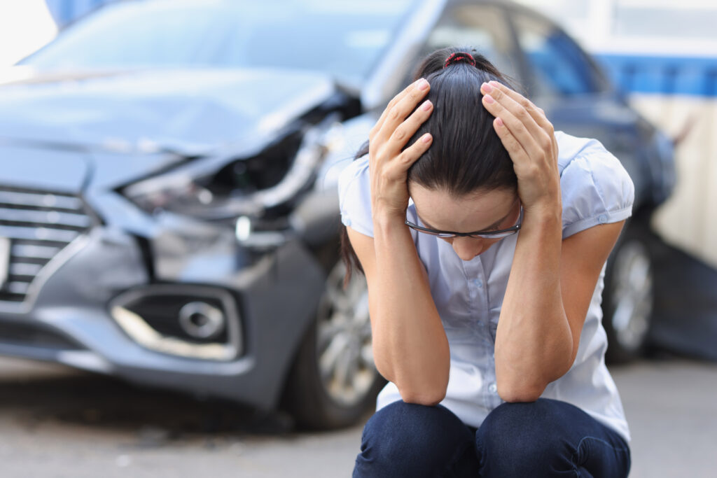 A young woman at a broken car sits holding her head